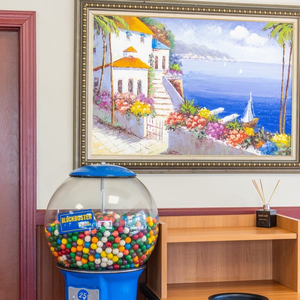 A gumball machine, a framed painting of a coastal town, and a wooden shelf with a diffuser.